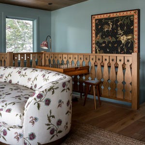 May include: A cozy interior featuring a floral-patterned sofa, wooden railing, and a framed artwork with deer and forest imagery. The room has a light blue wall, wooden floor, and a small wooden stool. A lamp with a patterned shade sits on the railing.
