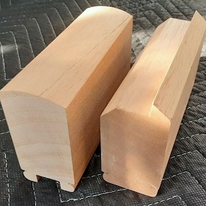 May include: Two rectangular blocks of light-colored, unfinished wood. The left block features a smooth, rounded top edge, while the right block has a beveled top edge. Both pieces have a distinct grooved profile along their bottom edges.