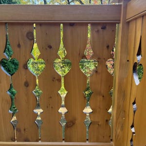 May include: Wooden deck railing with decorative cutouts in a repeating pattern. The cutouts are heart-shaped and diamond-shaped, with a natural wood finish. The railing is outdoors with a view of trees and sky.