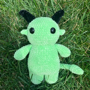 May include: A green crocheted plush toy with black horns and large black eyes. The toy is sitting on a bed of green grass.