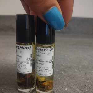 Puede incluir: Dos peque&ntilde;os frascos de vidrio con tapas negras y etiquetas. Las etiquetas son blancas con texto negro y un peque&ntilde;o gr&aacute;fico de flores. El texto en las etiquetas dice "t(Him) Gems Sint selt. sen you for me! 402.298.3810 Enhanced***" y "t(Her) Oil Gems Maha W Dreams.v to me! 402.298.1810 Maint selt. Alo to help WOHan you Enhanced***". Los frascos est&aacute;n llenos de un l&iacute;quido transparente y contienen peque&ntilde;os trozos de hierbas y flores secas.