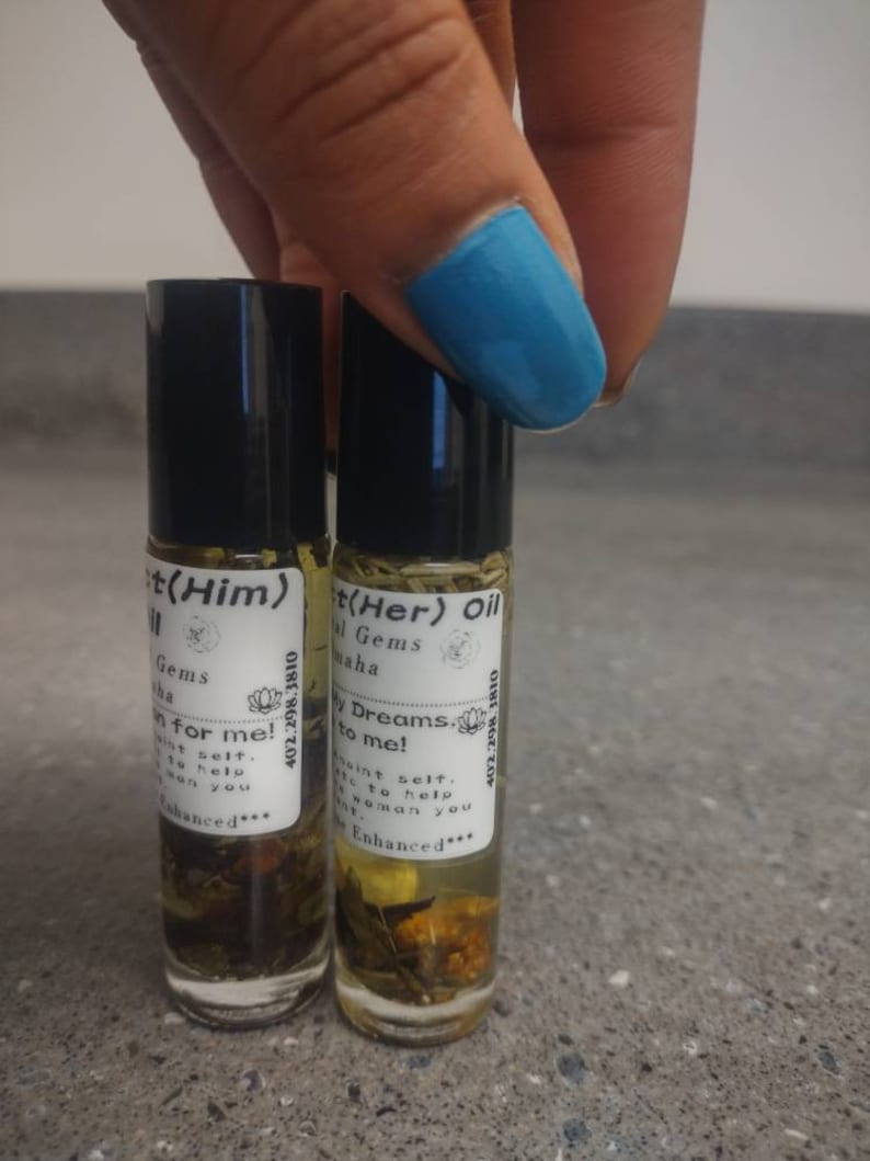 Puede incluir: Dos peque&ntilde;os frascos de vidrio con tapas negras y etiquetas. Las etiquetas son blancas con texto negro y un peque&ntilde;o gr&aacute;fico de flores. El texto en las etiquetas dice "t(Him) Gems Sint selt. sen you for me! 402.298.3810 Enhanced***" y "t(Her) Oil Gems Maha W Dreams.v to me! 402.298.1810 Maint selt. Alo to help WOHan you Enhanced***". Los frascos est&aacute;n llenos de un l&iacute;quido transparente y contienen peque&ntilde;os trozos de hierbas y flores secas.