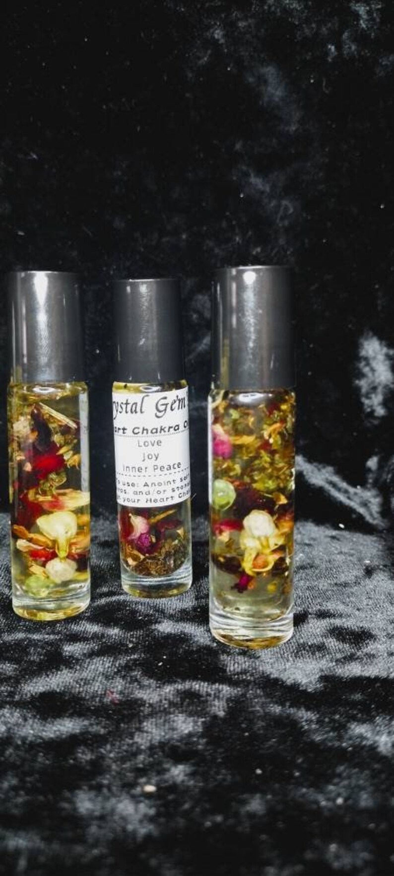 Puede incluir: Tres botellas de vidrio con rodillo llenas de un l&iacute;quido transparente y varias flores y hierbas secas. Las botellas est&aacute;n etiquetadas con "Crystal Gem's Heart Chakra Oil", "Love", "Joy", "Inner Peace", "Use: Anoint self and/or stones on your Heart Chakra"
