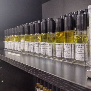 Puede incluir: Un estante lleno de peque&ntilde;as botellas de vidrio que contienen l&iacute;quido amarillo. Las botellas tienen tapas negras y etiquetas con texto que incluye "Woman in Oil", "Crystal Gems", "Healing Real" y "Bugs Be Gone".