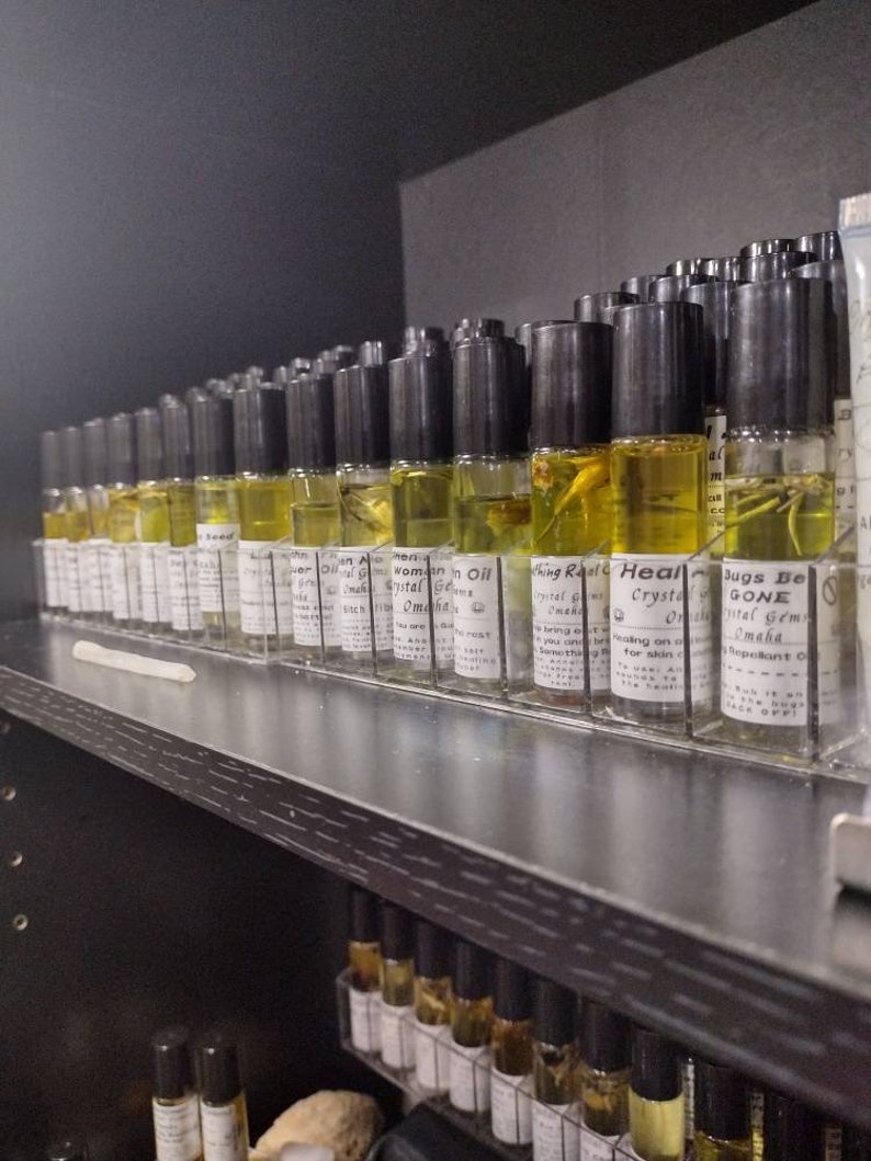 Puede incluir: Un estante lleno de peque&ntilde;as botellas de vidrio que contienen l&iacute;quido amarillo. Las botellas tienen tapas negras y etiquetas con texto que incluye "Woman in Oil", "Crystal Gems", "Healing Real" y "Bugs Be Gone".