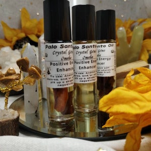 Puede incluir: Tres botellas de vidrio con aplicadores de rodillo llenas de un l&iacute;quido &aacute;mbar claro. Las botellas est&aacute;n etiquetadas con "Palo Santo Oil", "Crystal Gems Omaha" y "Positive Energy Enhancer". Las botellas est&aacute;n sobre una bandeja plateada con un pa&ntilde;o blanco debajo.