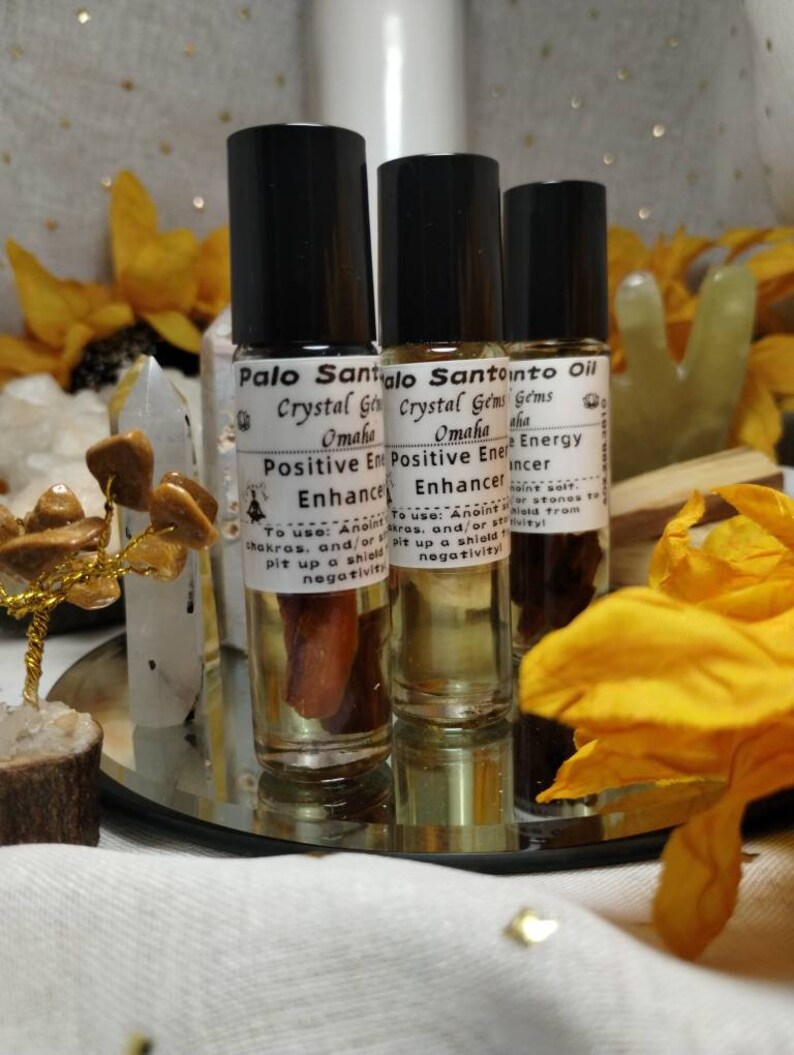 Puede incluir: Tres botellas de vidrio con aplicadores de rodillo llenas de un l&iacute;quido &aacute;mbar claro. Las botellas est&aacute;n etiquetadas con "Palo Santo Oil", "Crystal Gems Omaha" y "Positive Energy Enhancer". Las botellas est&aacute;n sobre una bandeja plateada con un pa&ntilde;o blanco debajo.