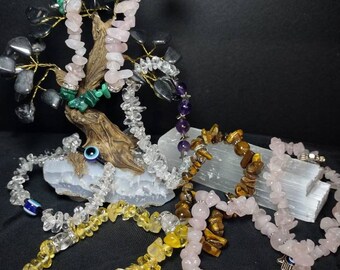 Puede incluir: Una colección de pulseras de virutas de piedras preciosas en varios colores, incluyendo amarillo, transparente, rosa y marrón. Una escultura de árbol de alambre con hojas de piedras preciosas se encuentra en el fondo. Las pulseras se exhiben sobre una superficie negra.