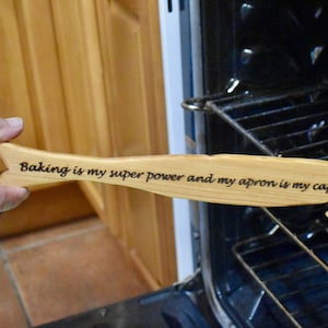 Puede incluir: Una espátula de madera con forma de pez con el texto "Baking is my super power and my apron is my cape." La espátula se sostiene frente a un horno abierto.