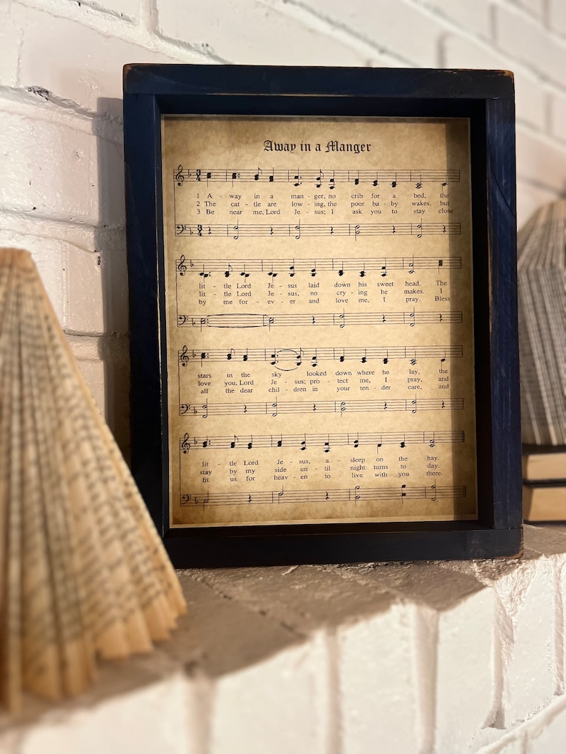 May include: A framed print of sheet music with the title "Away in a Manger". The music is printed in black and white on a cream-colored background. The frame is black.