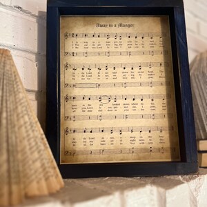 May include: A framed print of a vintage sheet music score for the Christmas carol "Away in a Manger". The sheet music is printed on aged paper with a brown tint and features musical notes and lyrics.