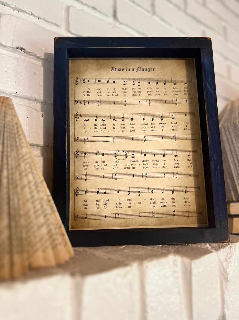 May include: A framed print of a vintage sheet music score for the Christmas carol "Away in a Manger". The sheet music is printed on aged paper with a brown tint and features musical notes and lyrics.