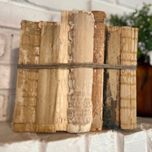 May include: A bundle of vintage-style books tied together with twine. The books have aged, textured covers in shades of beige and brown. The spines are visible, and the overall aesthetic is rustic and decorative.