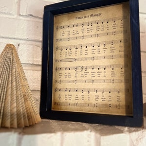 May include: A framed print of the song "Away in a Manger" with sheet music and lyrics. The print is on aged paper with a brown background and black text. The frame is dark blue.