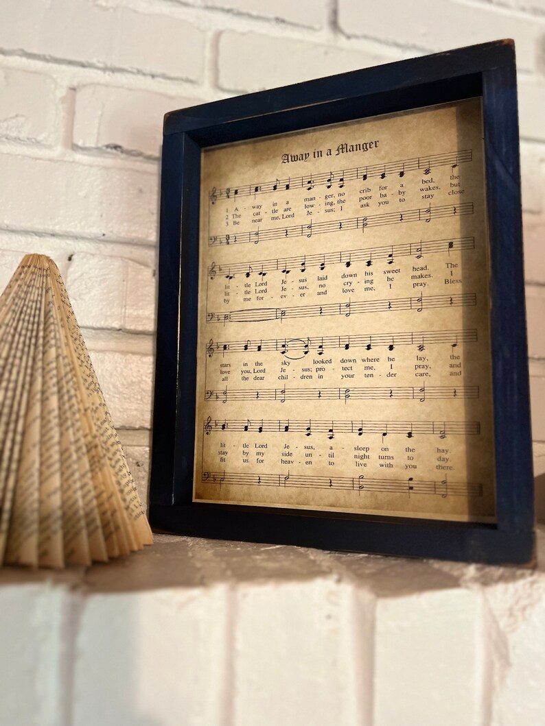 May include: A framed print of the song "Away in a Manger" with sheet music and lyrics. The print is on aged paper with a brown background and black text. The frame is dark blue.