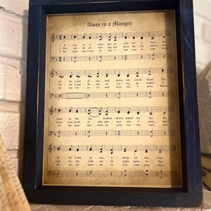 May include: A framed vintage sheet music print with the title "Away in a Manger". The lyrics and musical notes are printed on aged paper with a brown tint. The frame is dark blue.