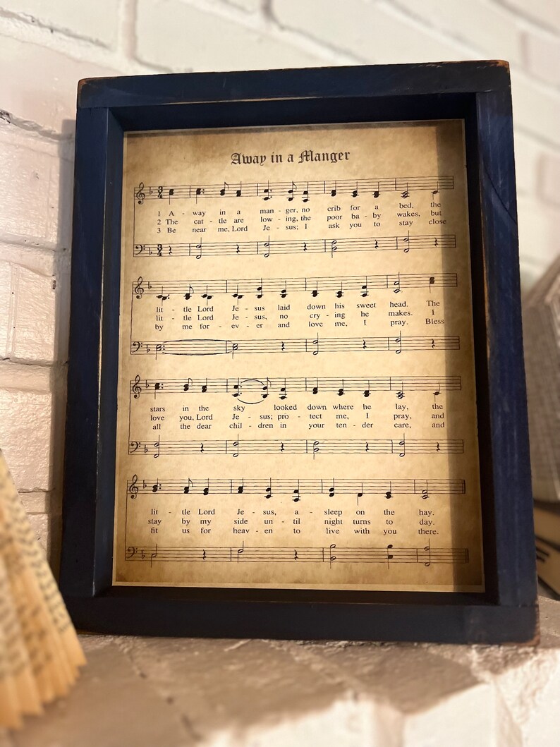 May include: A framed vintage sheet music print with the title "Away in a Manger". The lyrics and musical notes are printed on aged paper with a brown tint. The frame is dark blue.