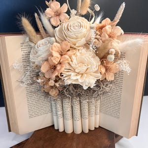 May include: A floral arrangement featuring cream and peach-colored flowers, wheat, and dried accents, set within an open vintage book. The base is composed of rolled book pages, creating a unique display.