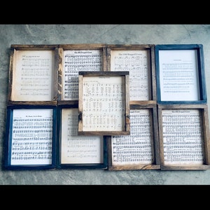 Puede incluir: Nueve piezas de m&uacute;sica en partituras enmarcadas con diferentes t&iacute;tulos de canciones, incluyendo "The Old Rugged Cross", "Amazing Grace" y "My Jesus, I Love Thee". Los marcos son de madera con un acabado r&uacute;stico y tienen un fondo negro o marr&oacute;n.