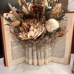 May include: A decorative floral arrangement featuring dried flowers and foliage in shades of brown, beige, and green. The arrangement is set in an open book with pages rolled to create a vase-like structure. The book has a red cover.