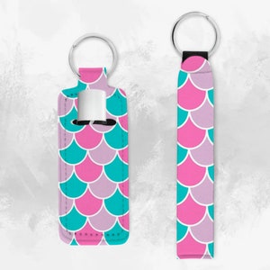May include: Two keychains with a mermaid scale pattern in pink, teal, and purple. One keychain is a loop with a strap and the other is a holder for a small bottle.