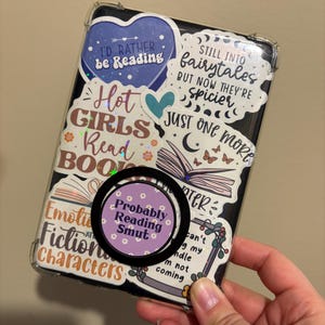 May include: A clear phone case with various colorful stickers featuring book-themed quotes and illustrations. The stickers include phrases like "I'd rather be reading", "Hot girls read books", "Still into fairytales but now they're spicier", and "Just one more chapter". A purple sticker with white flowers and the text "Probably reading smut" is attached to a black phone ring.