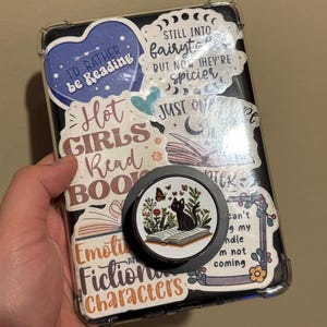 May include: A clear phone case with a variety of colorful stickers, including a black cat sitting on a book, a heart with the text "I'd rather be reading", and a moon with the text "Still into fairytales but now they're spicier".