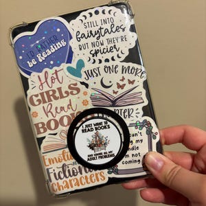 May include: A clear phone case with various colorful stickers featuring book-themed quotes and illustrations. The stickers include phrases like "I'd rather be reading", "Hot girls read books", "Still into fairytales but now they're spicier", and "Just one more chapter".