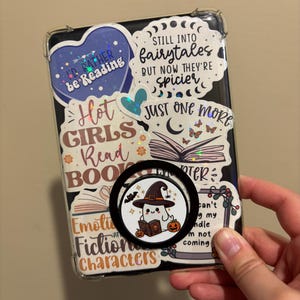 May include: A clear phone case with a variety of colorful stickers. The stickers feature quotes about reading, books, and fairytales. A black phone ring holder with a ghost wearing a witch hat and holding a book is attached to the back of the case.