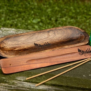 May include: A wooden incense holder with a carved design of trees and the text "Forestdale". The holder is made of dark wood and has a light brown finish. It is sitting on a wooden surface with two incense sticks and a green lighter with the text "Forestdale" on it.