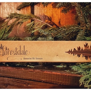 May include: A brown cardboard box with a cutout of a pine tree. The box contains incense sticks and has the text "Forestdale Essential Oil Incense" printed on the front.