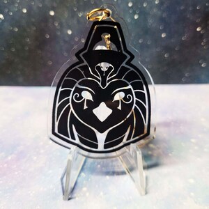 May include: Black and white acrylic keychain featuring a stylised depiction of the Egyptian god Horus. The keychain is on a clear acrylic stand.