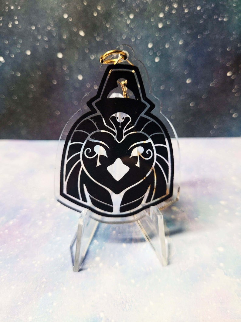 May include: Black and white acrylic keychain featuring a stylised depiction of the Egyptian god Horus. The keychain is on a clear acrylic stand.