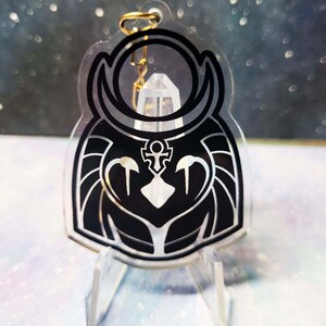 May include: Black and white acrylic keychain featuring a stylised owl design with a crescent moon and an ankh symbol. The keychain is on a clear acrylic stand.