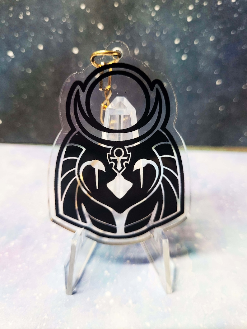 May include: Black and white acrylic keychain featuring a stylised owl design with a crescent moon and an ankh symbol. The keychain is on a clear acrylic stand.