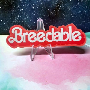 May include: A red sticker with white lettering that reads "Breedable". The sticker is on a clear plastic stand and is sitting on a light blue and pink watercolor background.