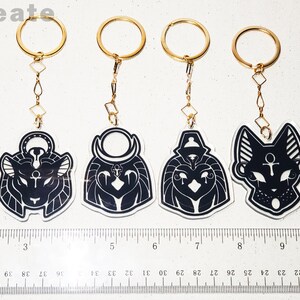 May include: Set of six black and white acrylic keychains featuring different Egyptian deity heads. Each keychain has a gold chain and ring. The keychains are approximately 5 cm tall.