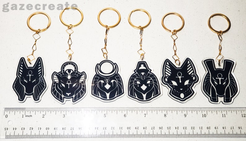 May include: Set of six black and white acrylic keychains featuring different Egyptian deity heads. Each keychain has a gold chain and ring. The keychains are approximately 5 cm tall.