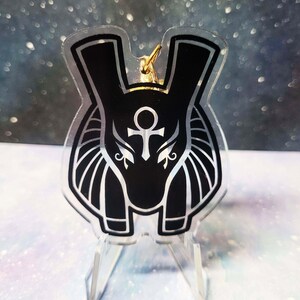 May include: Black and white acrylic keychain featuring an Anubis head with a gold Ankh symbol. The keychain is on a clear acrylic stand.
