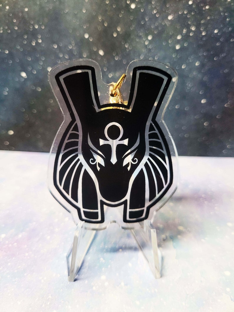 May include: Black and white acrylic keychain featuring an Anubis head with a gold Ankh symbol. The keychain is on a clear acrylic stand.