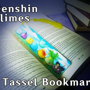 Puede incluir: Un marcapáginas de borla azul y amarillo con una variedad de slimes de Genshin Impact de colores. El marcapáginas tiene el texto "Genshin Slimes" en la parte superior y "Tassel Bookmark" en la parte inferior.