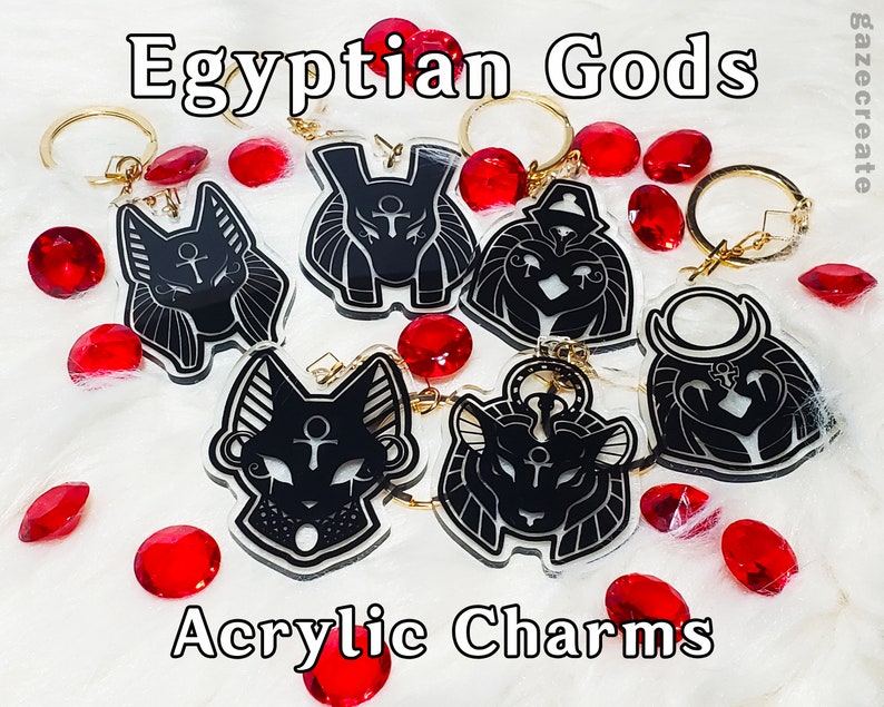 May include: Six black and white acrylic charms featuring Egyptian gods. The charms are attached to gold key rings. The charms are on a white surface with red gems scattered around them. The text "Egyptian Gods Acrylic Charms" is at the bottom of the image.