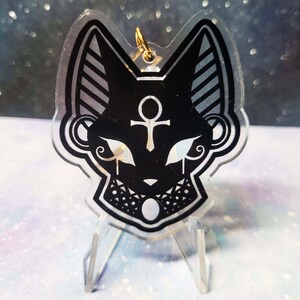 May include: Black and white acrylic charm featuring a stylised cat head with an ankh symbol and a crescent moon. The charm is on a clear acrylic stand.