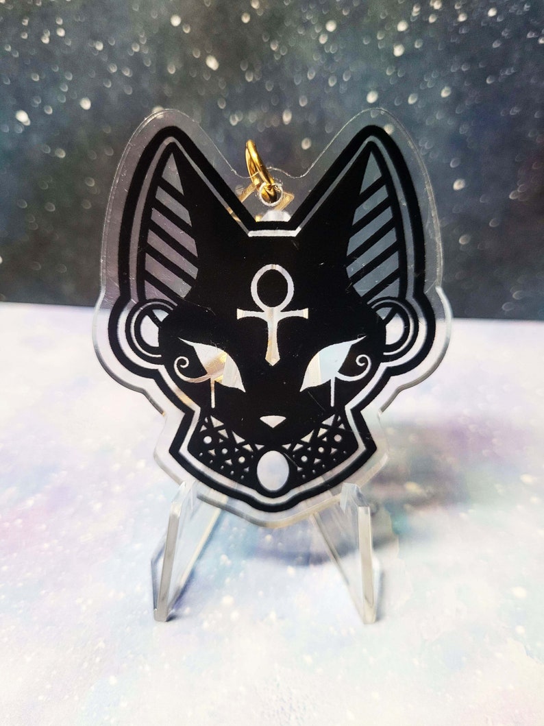 May include: Black and white acrylic charm featuring a stylised cat head with an ankh symbol and a crescent moon. The charm is on a clear acrylic stand.