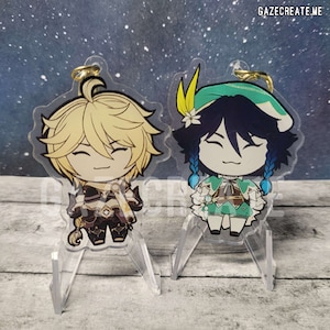Puede incluir: Dos charms de personajes de estilo anime en acrílico, uno con cabello rubio y un atuendo negro y dorado, el otro con cabello azul y un atuendo verde y blanco. Ambos charms están en soportes de acrílico transparente.