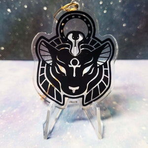 May include: A black and gold acrylic charm featuring a stylised depiction of an Egyptian deity with a cat head, a golden ankh symbol, and a golden halo. The charm is on a clear acrylic stand.