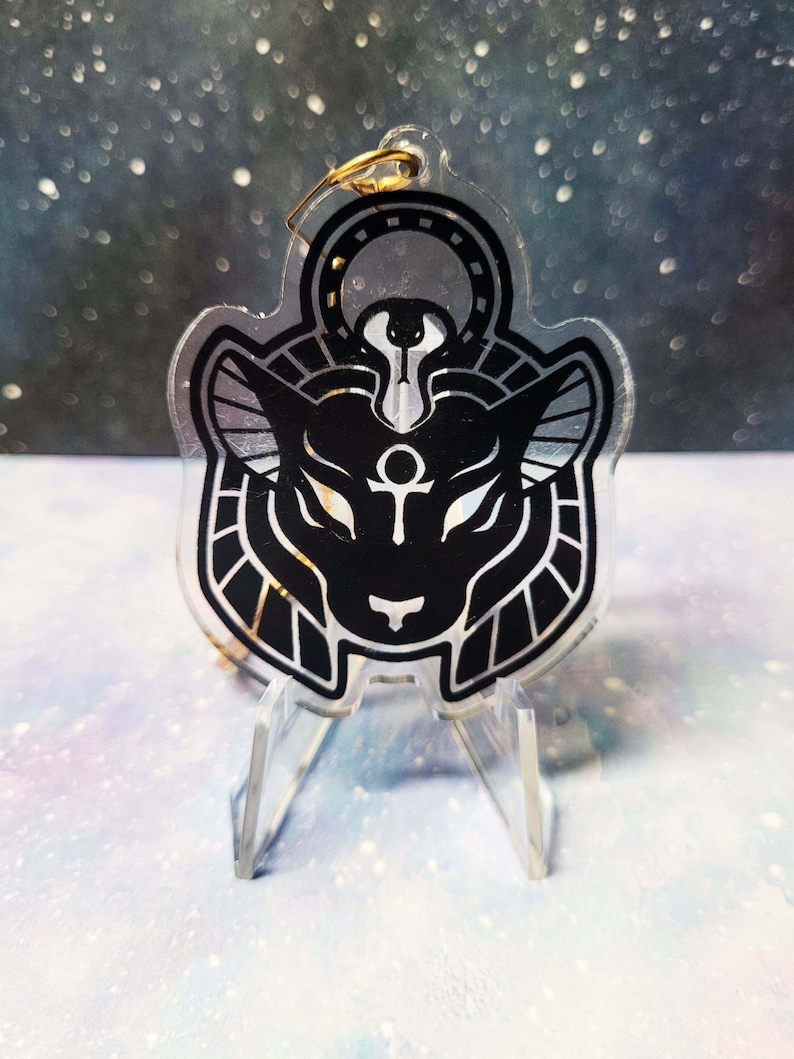 May include: A black and gold acrylic charm featuring a stylised depiction of an Egyptian deity with a cat head, a golden ankh symbol, and a golden halo. The charm is on a clear acrylic stand.
