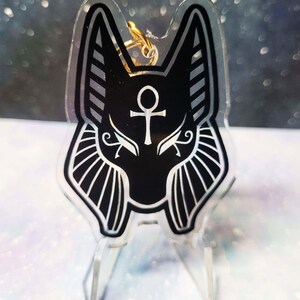 May include: Black and white acrylic keychain featuring an Anubis jackal head with a gold ankh symbol on its forehead. The keychain is on a clear acrylic stand.