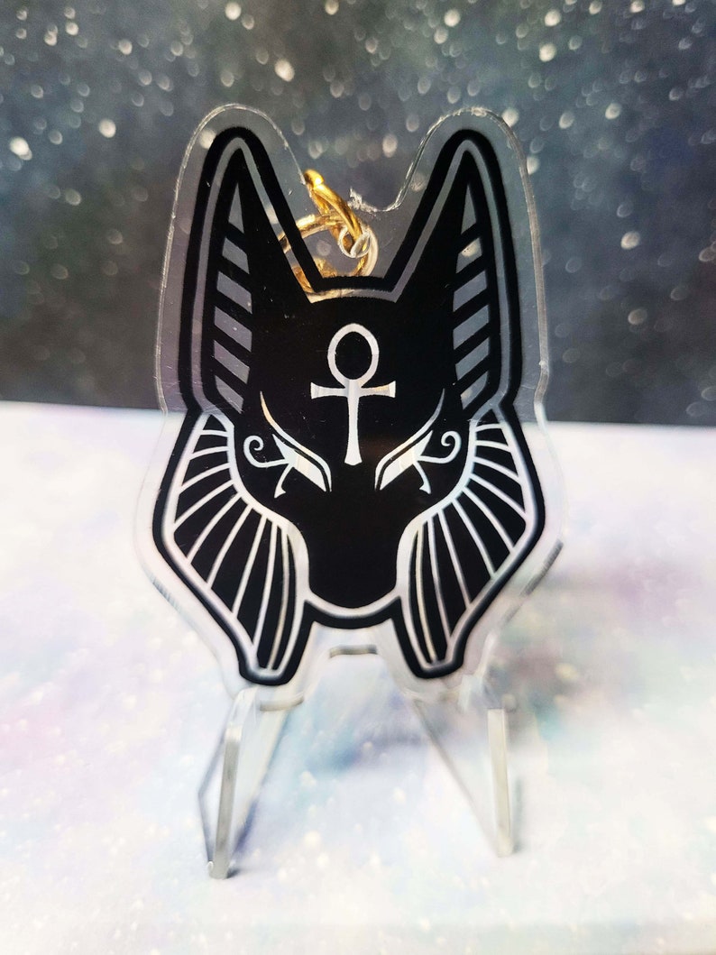 May include: Black and white acrylic keychain featuring an Anubis jackal head with a gold ankh symbol on its forehead. The keychain is on a clear acrylic stand.