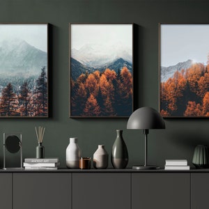 May include: Three framed landscape photographs of mountains and trees with fall foliage. The photos are hung on a dark green wall above a shelf with decorative objects.